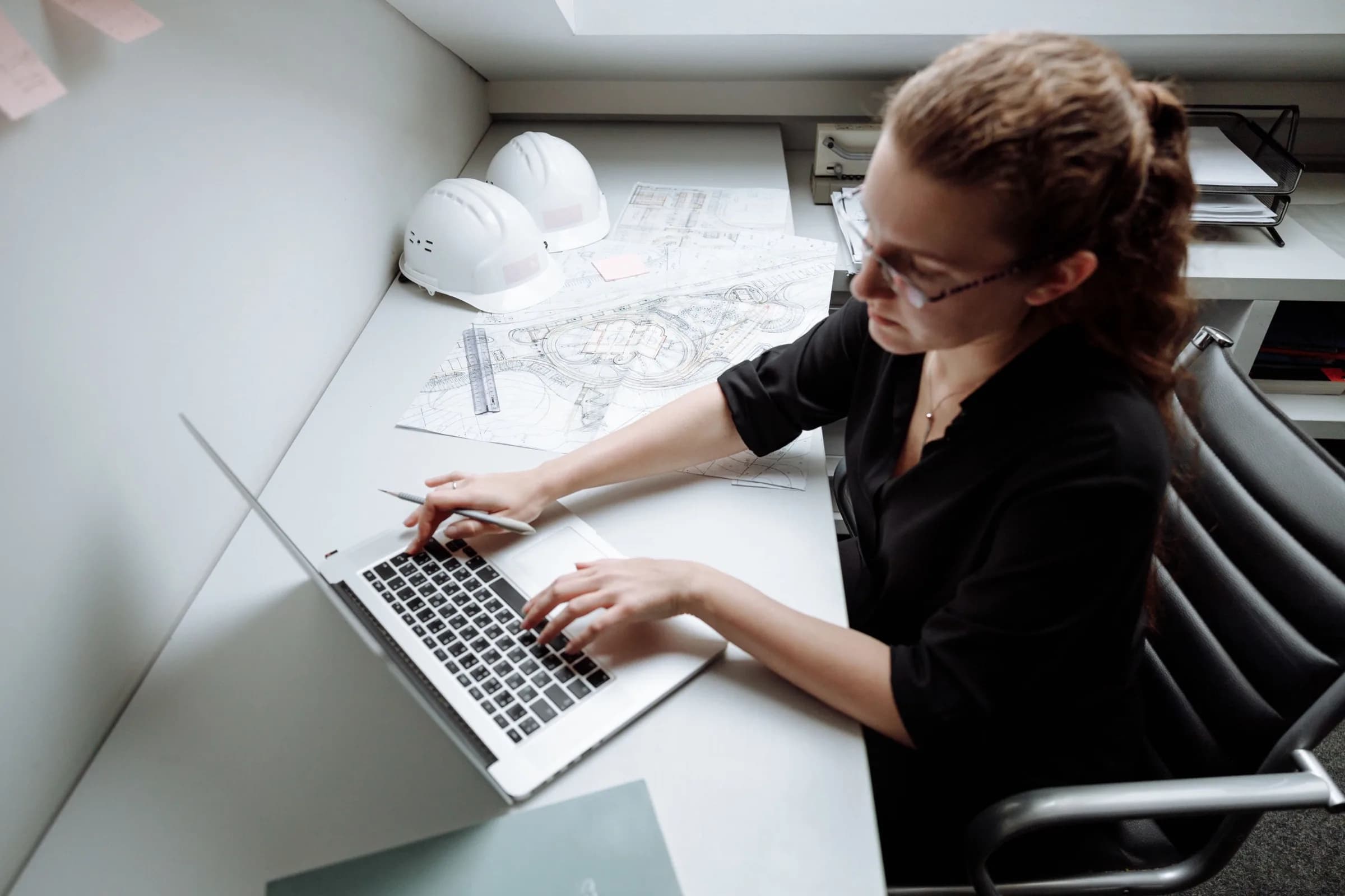 An estimator working on a laptop at a desk, a printed drawing set open beside it and a hard hat within reach