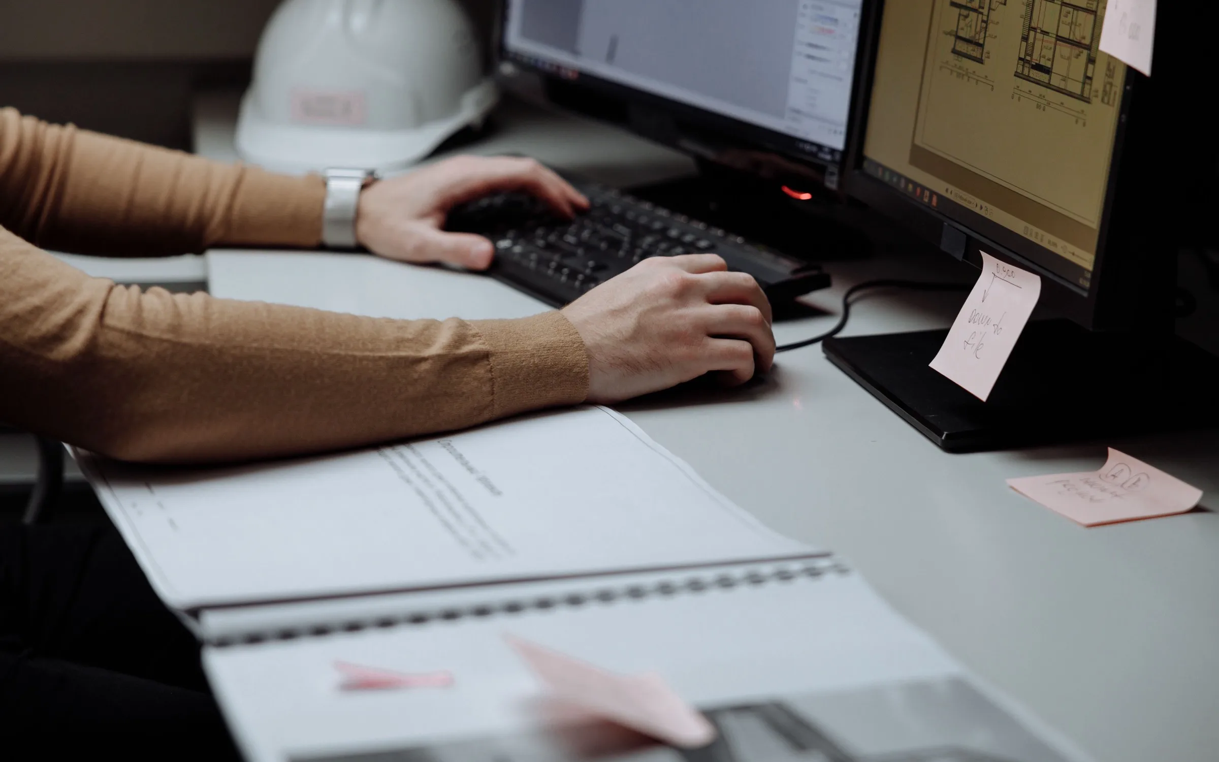 An estimator working at two monitors with construction drawings on screen and a hard hat on the desk