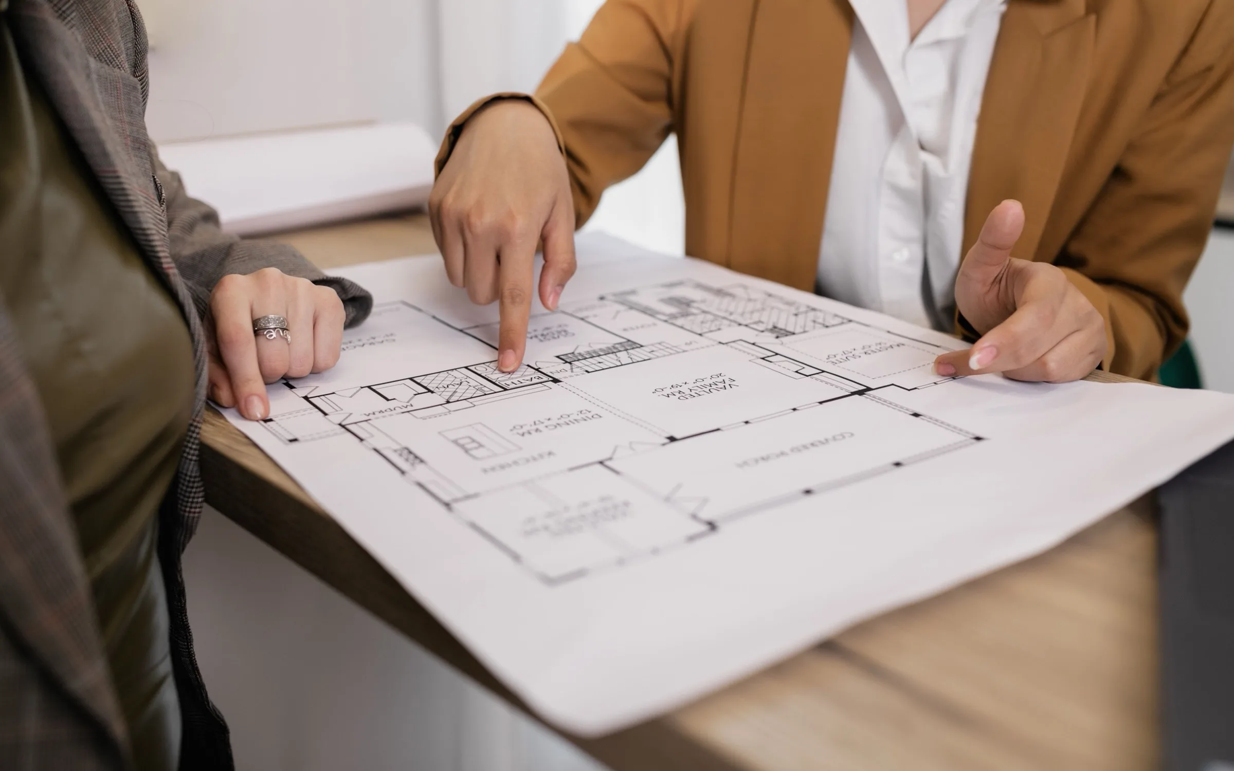 Two people reviewing a house floor plan together at a table, pointing at a room on the sheet